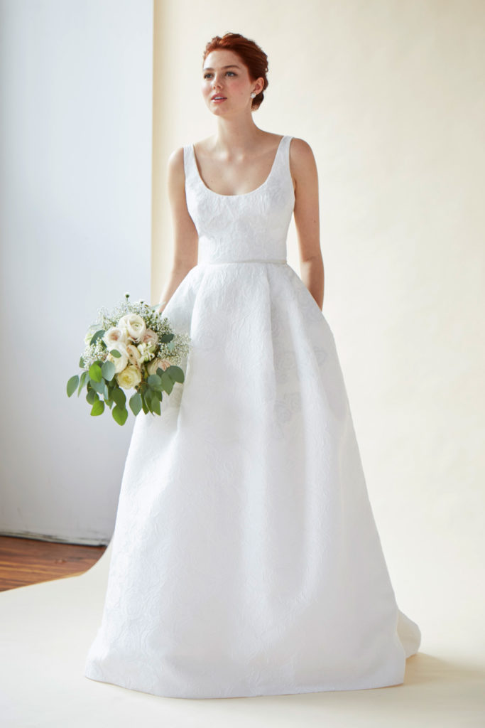 Bride wearing a classic scoop neck wedding dress with a full pleated skirt, standing against a bright white backdrop with a bouquet of white roses.
