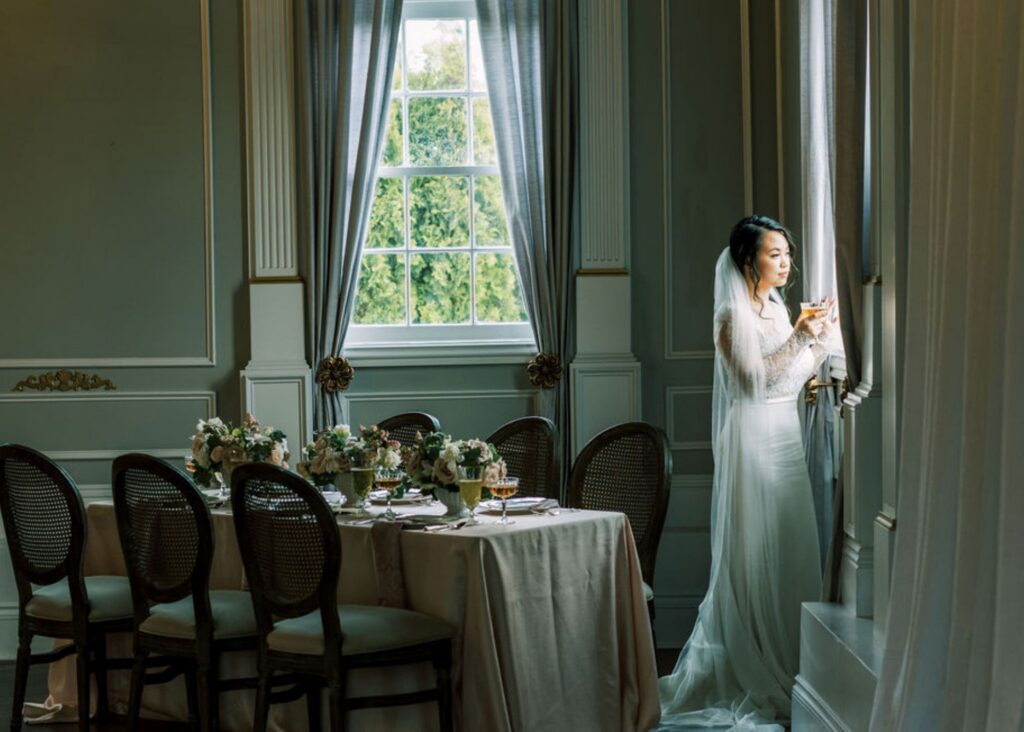 a bride wearing a vintage-inspired, sleek wedding dress with lace looks contemplatively out the window of a drawing room