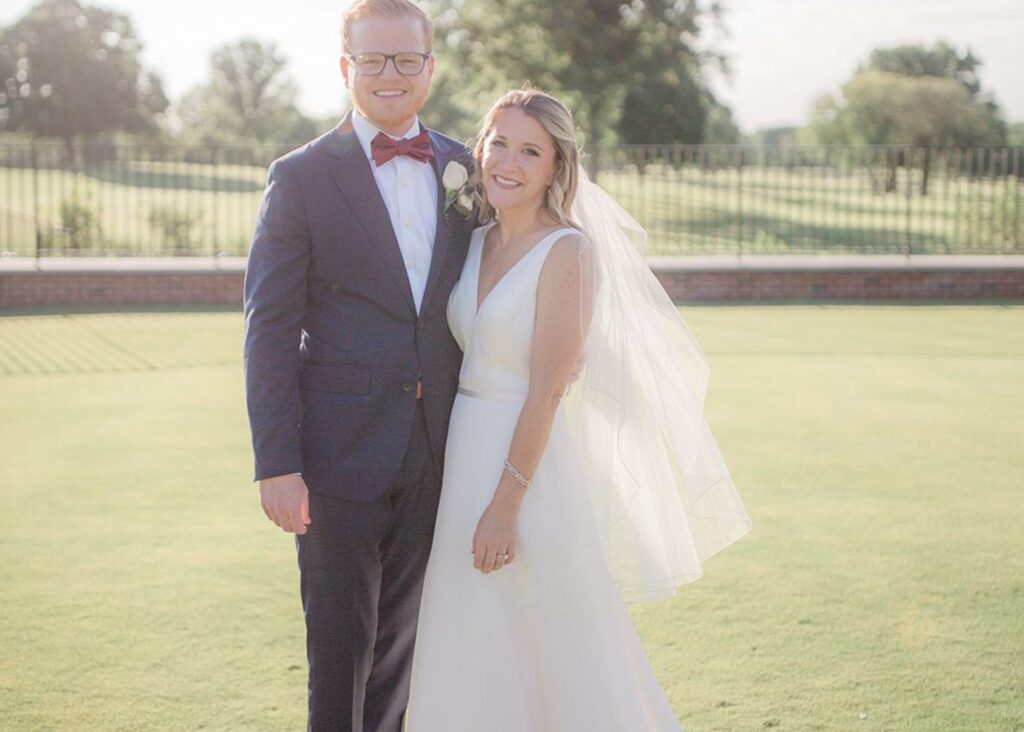 A bride and room stand in the sunshine smiling, the groom in a suit and the bride in a form fitting v-neck gown and fingertip veil