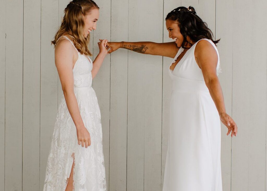 two brides hold hands and smile at each other during their first look. Both wear sleek wedding dresses.