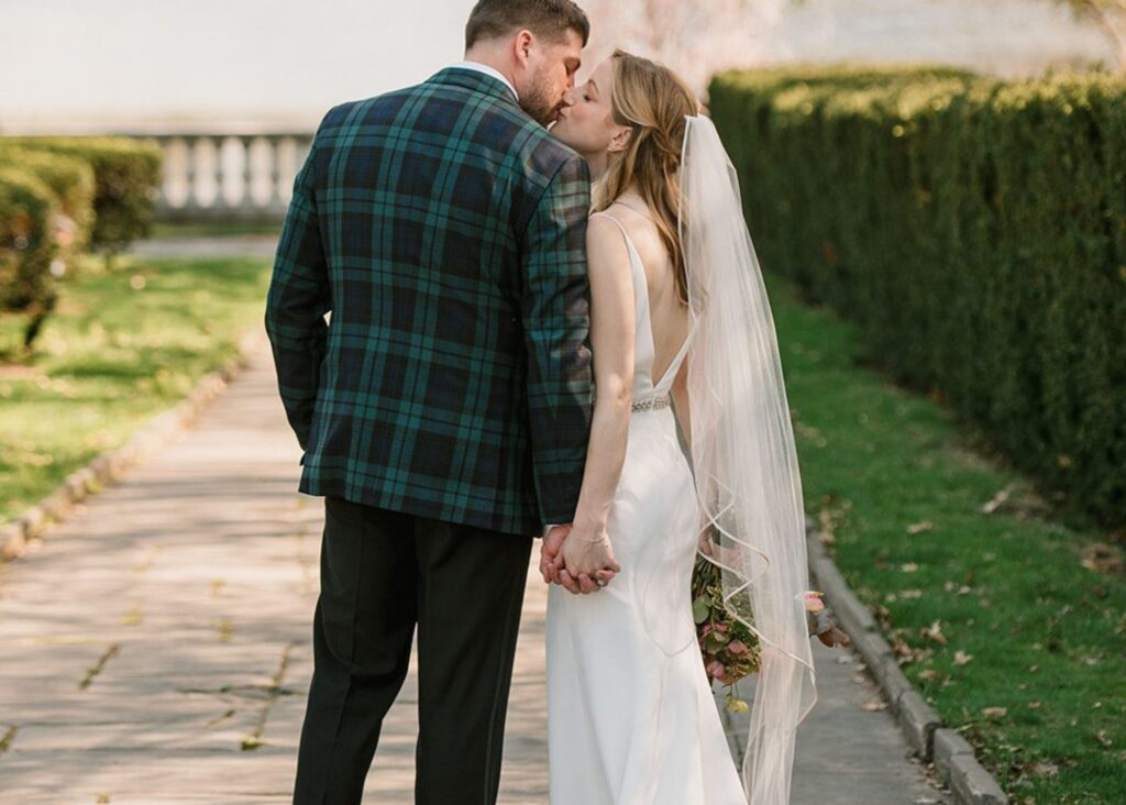 a groom wearing a tartan jacket kisses his bride in a sleek wedding dress and cathedral veil