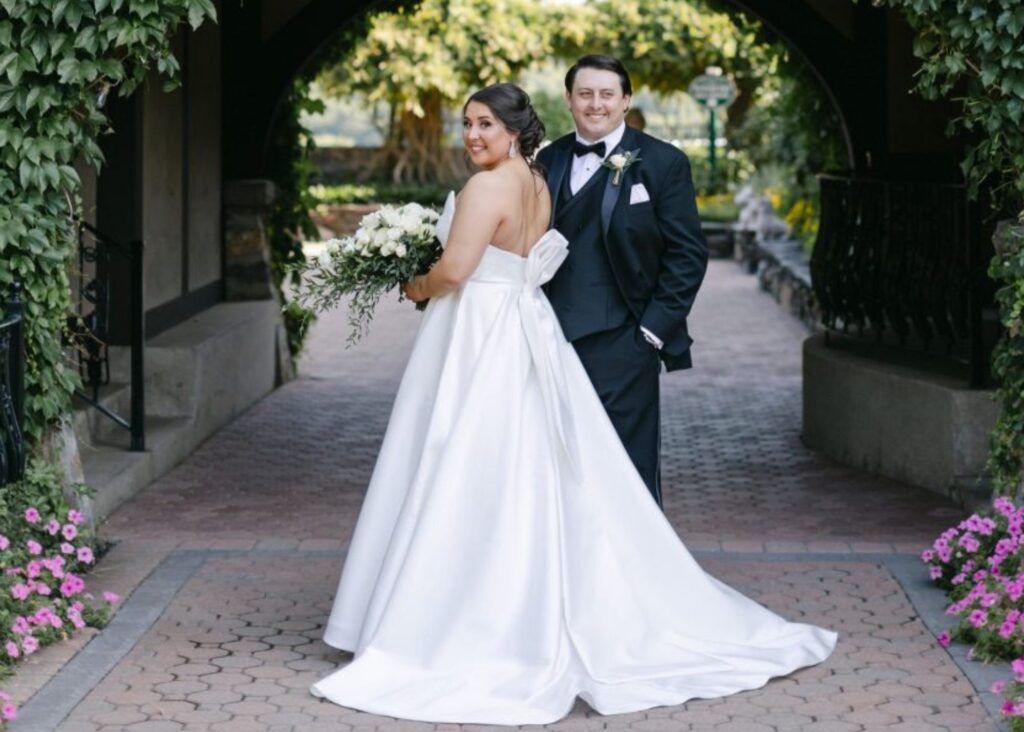 a bride and groom stand beneath a brick archway, the bride's back is to us and she smiles over her shoulder. She is wearing a ball gown with long wide tails.