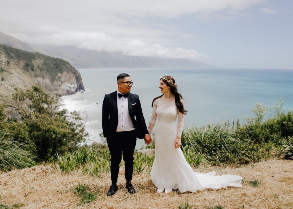 a bride and groom stand on a cliff in Big Sure, holding hands. The bride wears a flower crown and lace and crepe sleek wedding gown with her long hair down