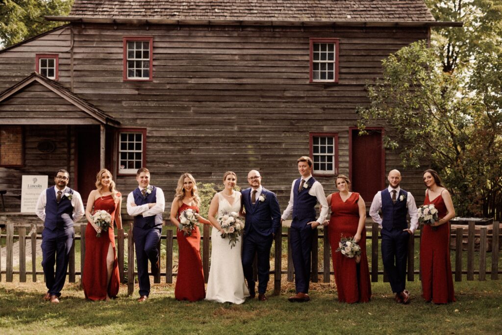A wedding party dressed in suits and crimson dresses stand in the sunlight. The bride wears a sleek wedding dress.