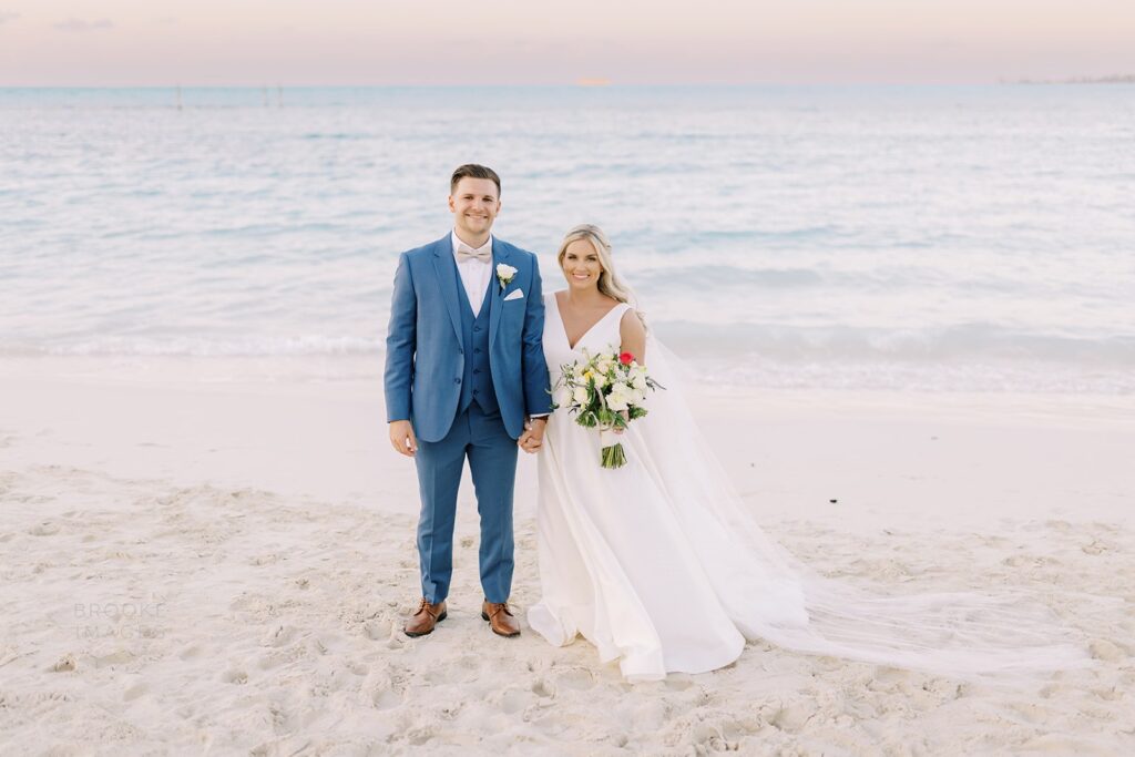 A bride and groom stand at sunset on a tropical beach. The bride wears a custom a-line wedding dress by Lea-Ann Belter bridal Toronto