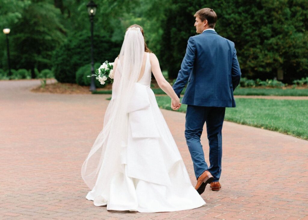 a bride and groom walk away hand in hand, the back gown of her A-line gown on display