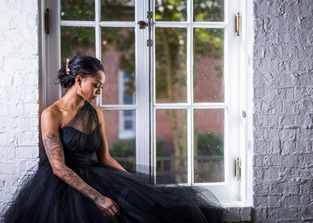 A woman wearing a black wedding dress with asymmetrical neckline sits in a window