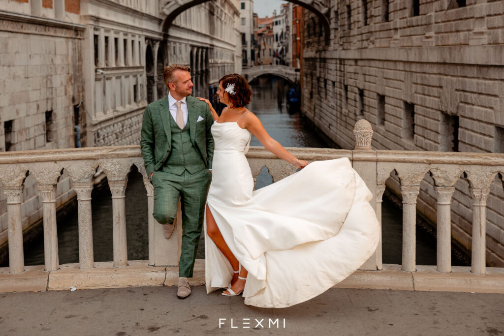 A bride and groom pose pin a bridge in Venice, Italy. The bride wears a sleek wedding dress by Lea-Ann Belter bridal.