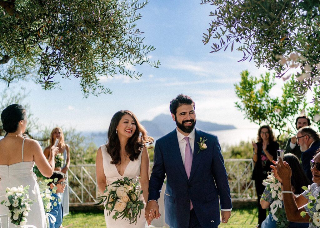 a bride and groom joyfully walk down the aisle in the Italian sunshine. The bride wears a sleek wedding dress, the groom a blue sit.
