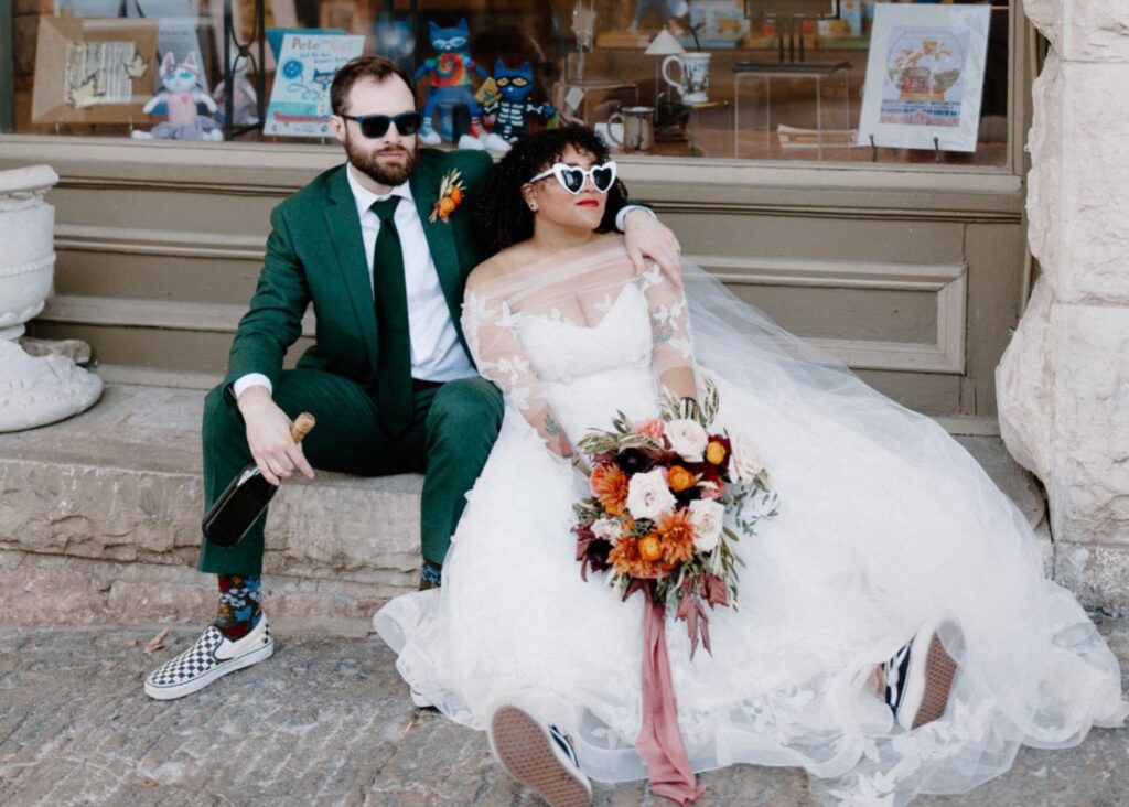 A bride and groom sit on a stoop wearing sunglasses. The bride wears her lace and net wedding gown and converse