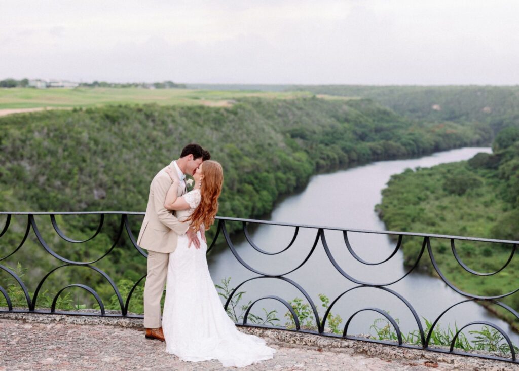 A bride and groom kiss passionately overlooking a river in the Dominican Republic. The bride wears her hair down and a lace fitted wedding dress