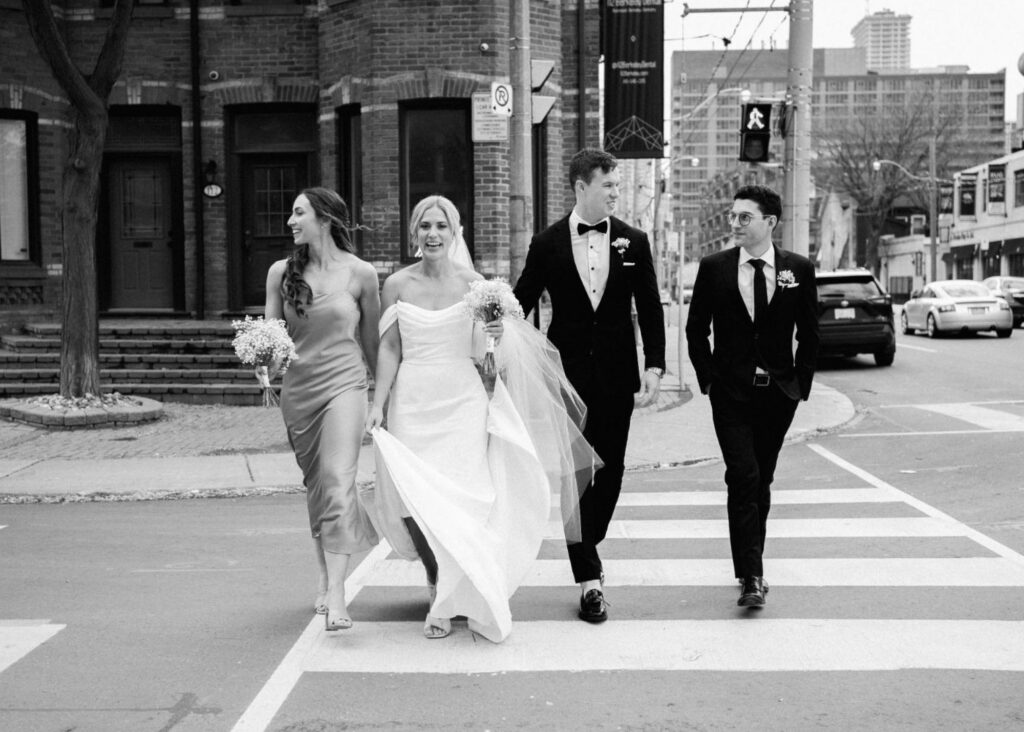 A black and white photo of a bride and groom and their wedding party on a Toronto cross walk. The bride wears an off the shoulder fitted dress, the groom a black tuyx