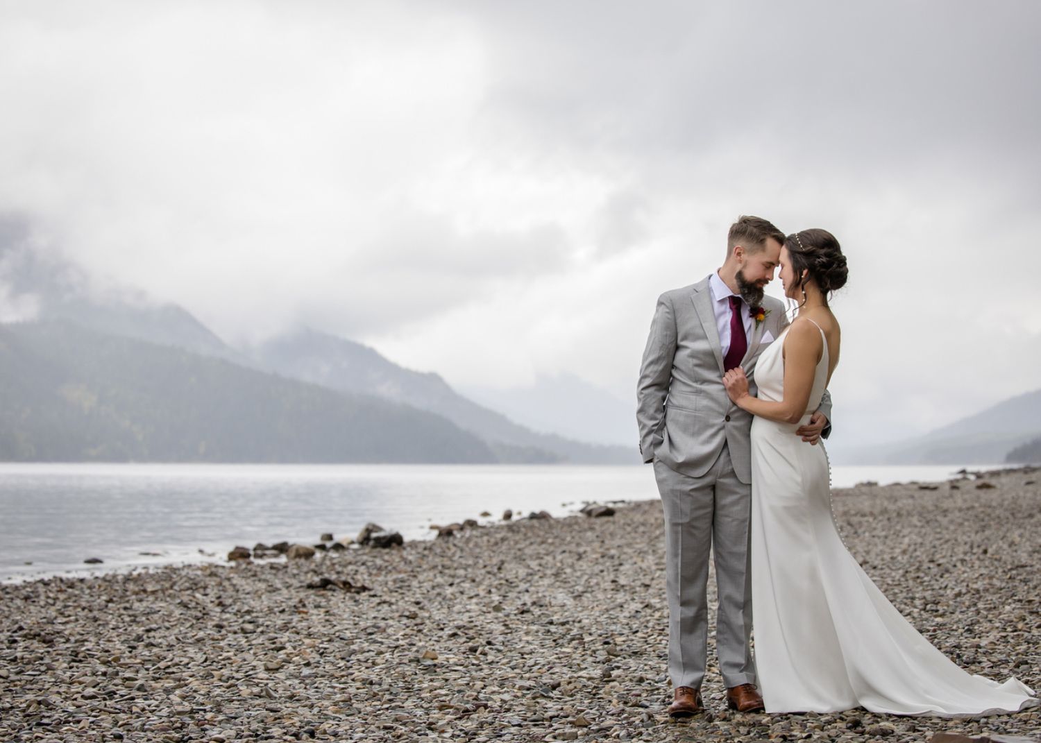 Joelle and Mathew stand in each others arms by the water in Waterton Park. Joelle is wearing a sleek wedding dress by Lea-Ann Belter Bridal