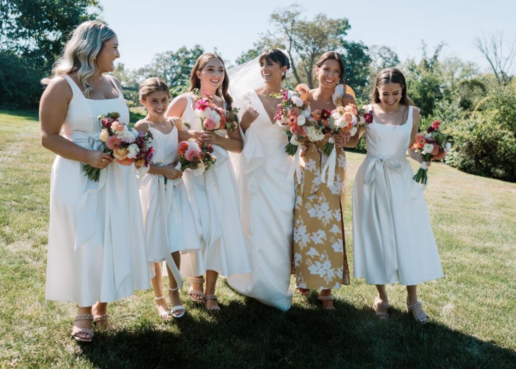 a bride weraing a sleek v-neck wedding dress walks with her bridesmaids in white across a sunny green meadow
