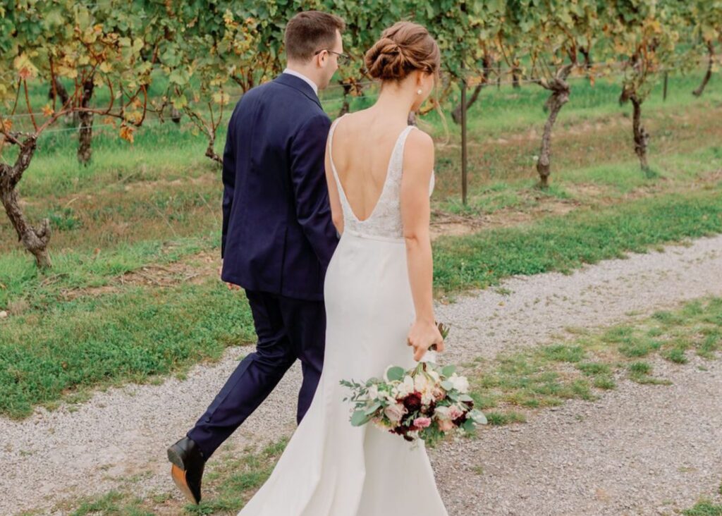 A bride adn groom walk away through a vineyard, the bride wears a low back sleek wedding dress