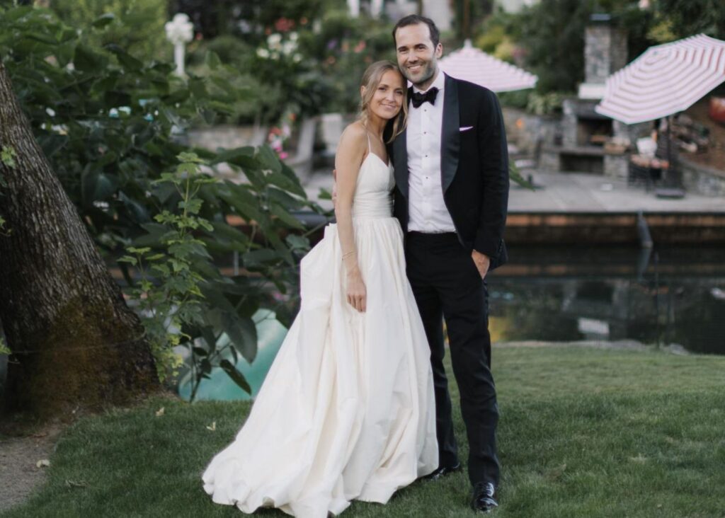 A brida and groom stand in each other's arms in a green garden under a tree. The bride wearins a ball gown wedding dress with camisole neckline.