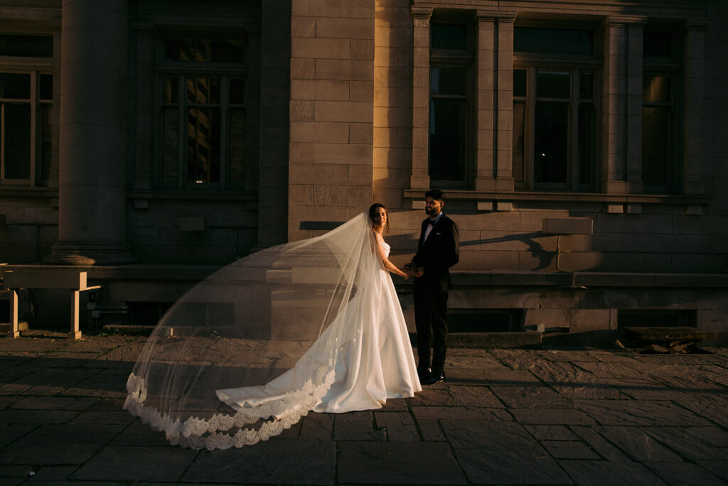 Bride and groom holding hands in golden hour light, bride wearing ballgown wedding dress with cathedral length lace veil