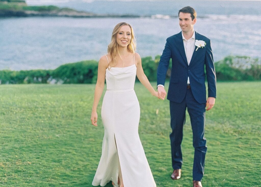 A bride and groom walk holding hands on the grass with the pacific ocean behind them. The bride wears a sleek wedding dress with slit, the groom a suit.