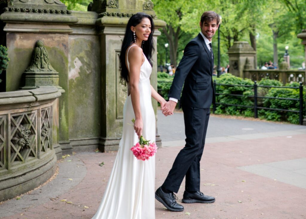 A bride and groom stand on a stone bridge in Central park. The bride wears a sleek, form fitting wedding dress