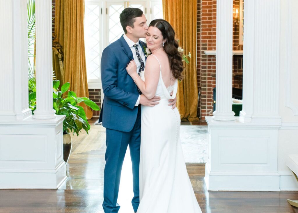 A groom holds his bride in an elegant drawing room. The bride wars a sleek crepe wedding dress by Lea-Ann Belter Bridal