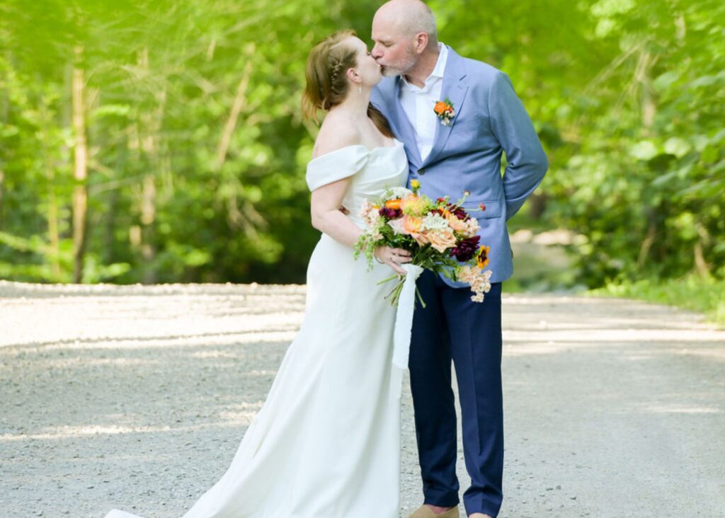 a bride wearing a sleek wedding dress and holding her sunflower bouquet kisses her groom in the woods.