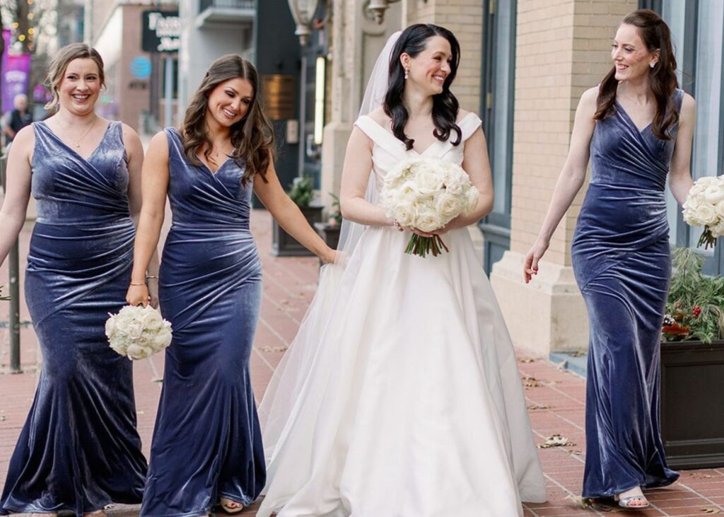 A bride in an off-the-shoudler ball gown walks with her bridesmaids in blue velvet in th sunshine