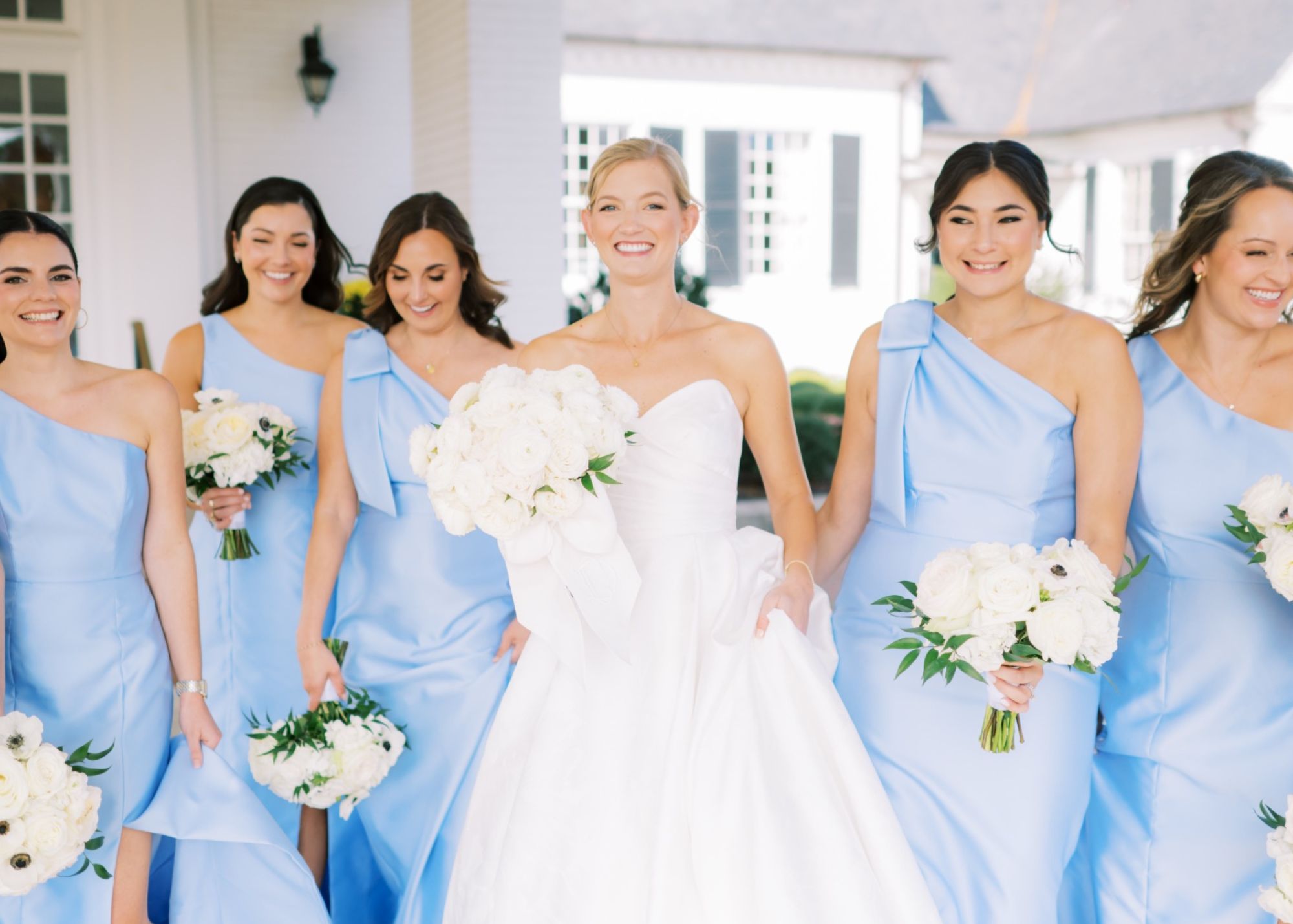 A beaming bride wearing a modern sweetheart ballgown wedding dress walks toward the camera with her bridesmaids wearing sky blue bridesmaids dresses