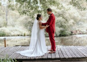 A bride stands in her groom's arms on a dock. The grom wears a red suit and the bride a sleek wedding dress with cathedral veil