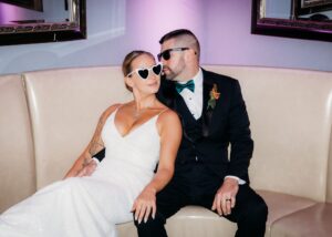 a bride and groom wearing white sunglasses kiss on a white banquette. She's wearing a sleek wedding dress with a v-neckline, he a tux.