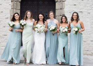 a bride wearing a sleek beaded wedding dress walks arm in arm with her bridesmaids in flowing gowns in shades of sea-glass blue in front of a stone church