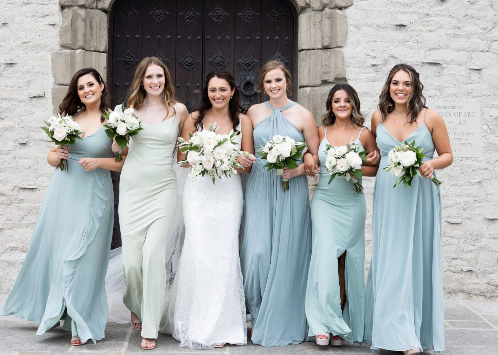 a bride wearing a sleek beaded wedding dress walks arm in arm with her bridesmaids in flowing gowns in shades of sea-glass blue in front of a stone church