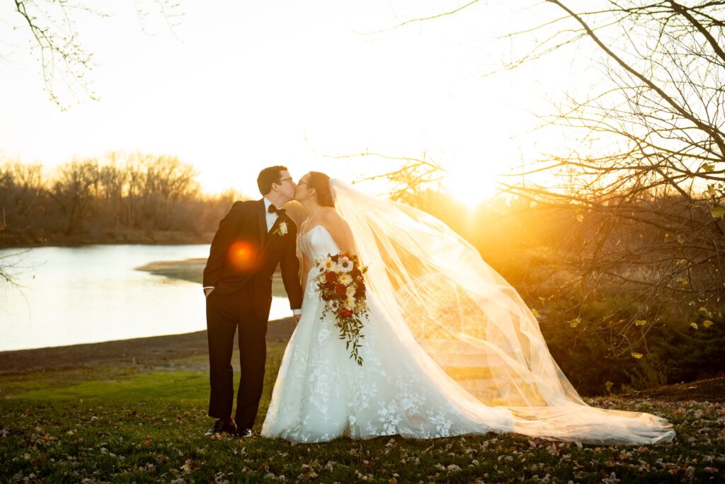 a bride wearing a gorgeous ball gown wedding dress with strapless neckline and A-line skirt kisses her groom before the sunset as her long cathedral veil flies behind her.