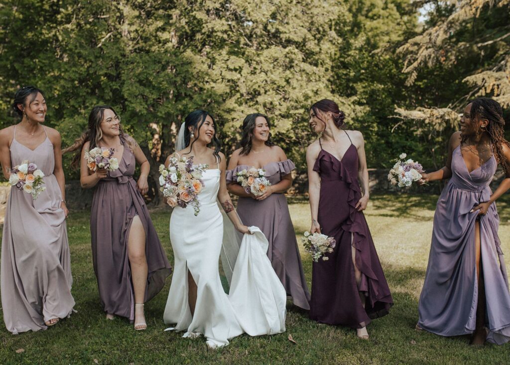 A bride and her wedding party walk toward the camera, the bridesmaids are wearing long gowns in purples and reds, and the bride wears a sleek, form fitting wedding dress
