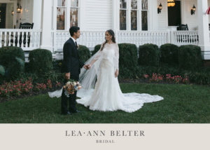 A bride and groom stand in a garden holding hands. The bride is wearing a lave wedding dress with a flowing lace cape