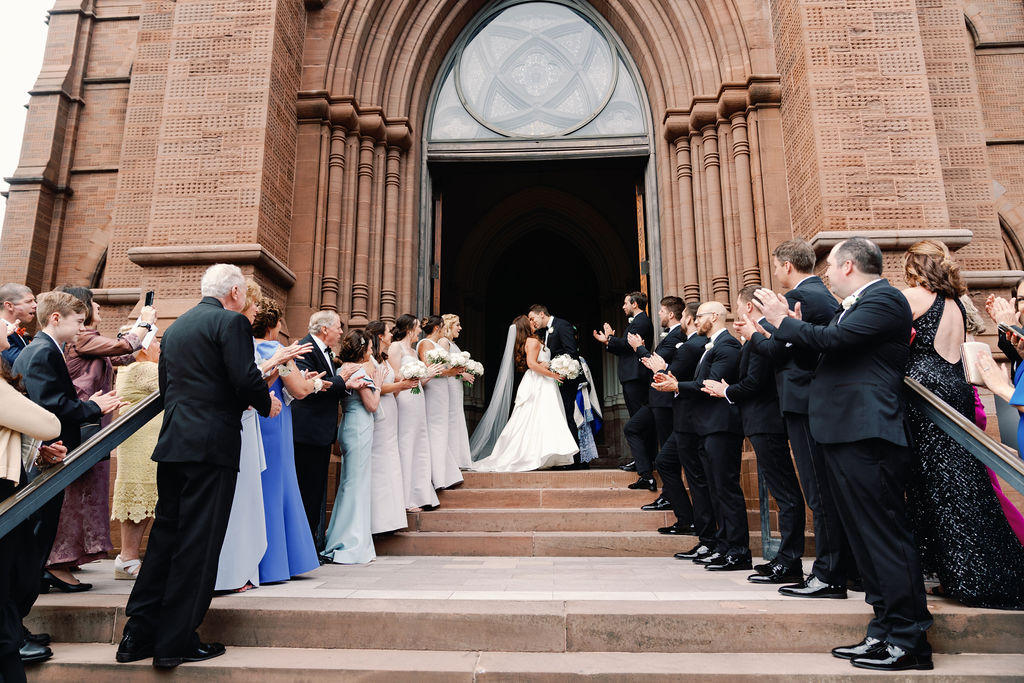 Bride and groom exiting church in ball gown wedding dress with long train as guests applaud on church steps