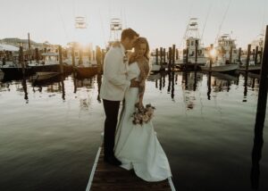 A bride and froom stand holding one another on a jetty in the early evening sunlight. The bride wears a strapless fit and flare wedding dress. The groom wears a white tuxedo jacket.