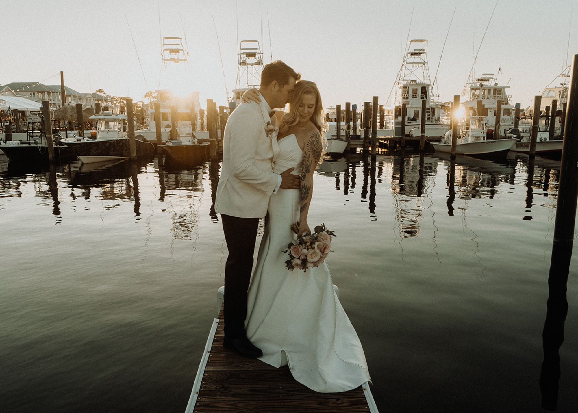 A bride and froom stand holding one another on a jetty in the early evening sunlight. The bride wears a strapless fit and flare wedding dress. The groom wears a white tuxedo jacket.