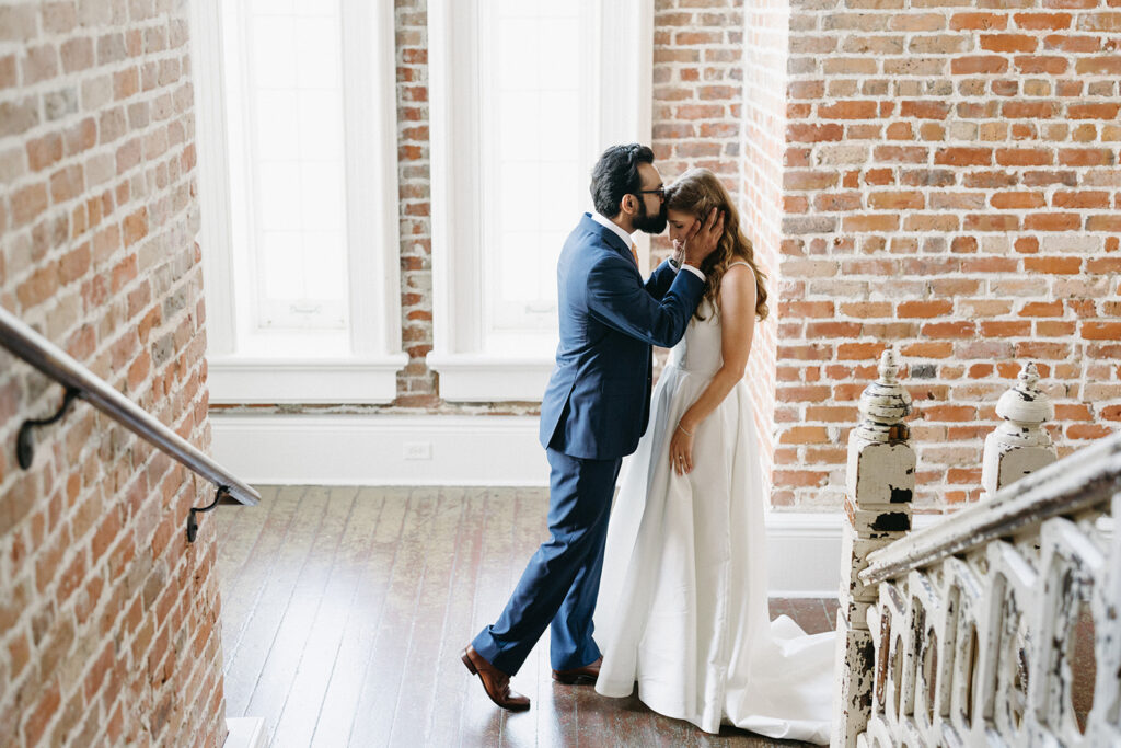 A groom kisses his bride in an exposed brick stairwell drenched in sunlight. The bride wears a classic bridal ball gown.