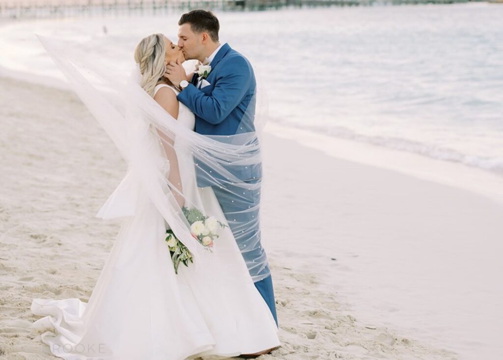 A bride and groom kiss on a Bahamas beach. The bride's pearl-dotted veil and A-line wedding dress swirl around her.