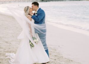 A bride and groom kiss on a Bahamas beach. The bride's pearl-dotted veil and A-line wedding dress swirl around her.