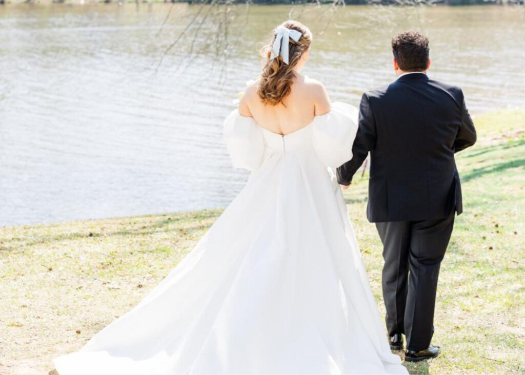 A bride and grom walk hand in hand o a sunny day beside a lake, The bride's A-line wedding dress billows out behind her.