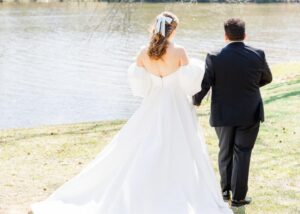 A bride and grom walk hand in hand o a sunny day beside a lake, The bride's A-line wedding dress billows out behind her.