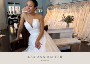 Smiling bride in a strapless wedding gown with a pointed sweetheart neckline and floral appliqué, at the Lea-Ann Belter Toronto bridal shop surrounded by racks of gowns
