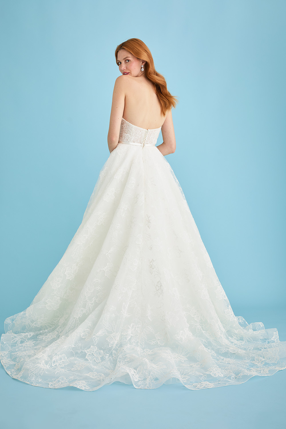 Bride wearing the Lea-Ann Belter Bridal Jane lace wedding dress, shown from behind to highlight the open back, satin waistband, and full circle skirt with a flowing lace train. The gown features delicate corded and Chantilly lace over silk organza and tulle.