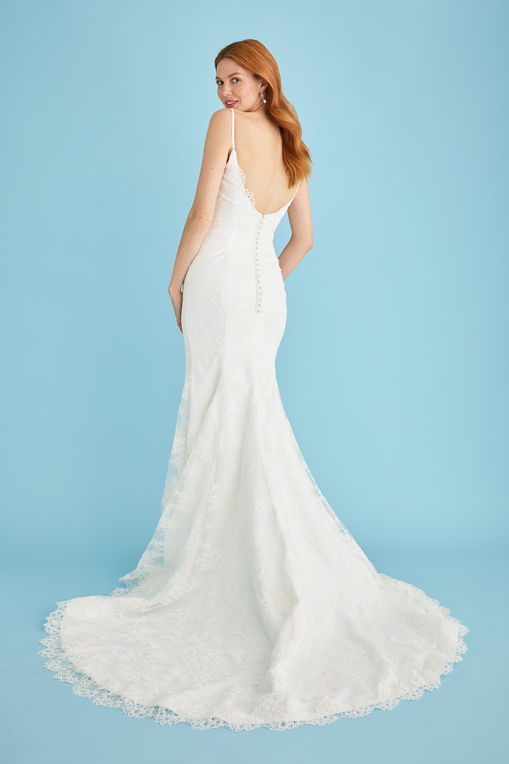 Bride wearing the Lea-Ann Belter Bridal Kitty fit-and-flare wedding dress, shown from the back to highlight the deep open back, scalloped Chantilly lace edging, and elegant sweep train. The gown features spaghetti straps and fabric-covered buttons.