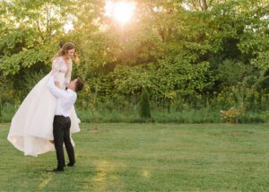 A goom lifts his bride above his head, the sunlight golden all around them. The bride wears a lace-topped gown with flowing organza bottom