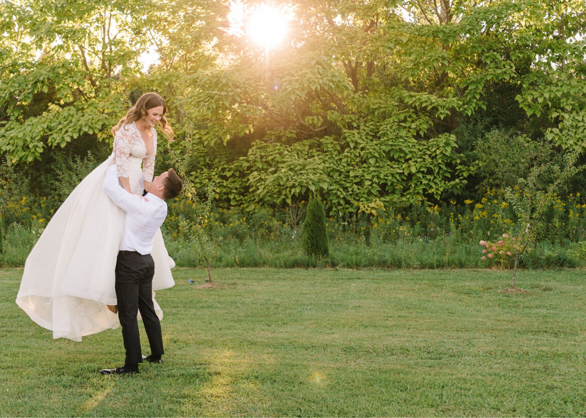 A goom lifts his bride above his head, the sunlight golden all around them. The bride wears a lace-topped gown with flowing organza bottom