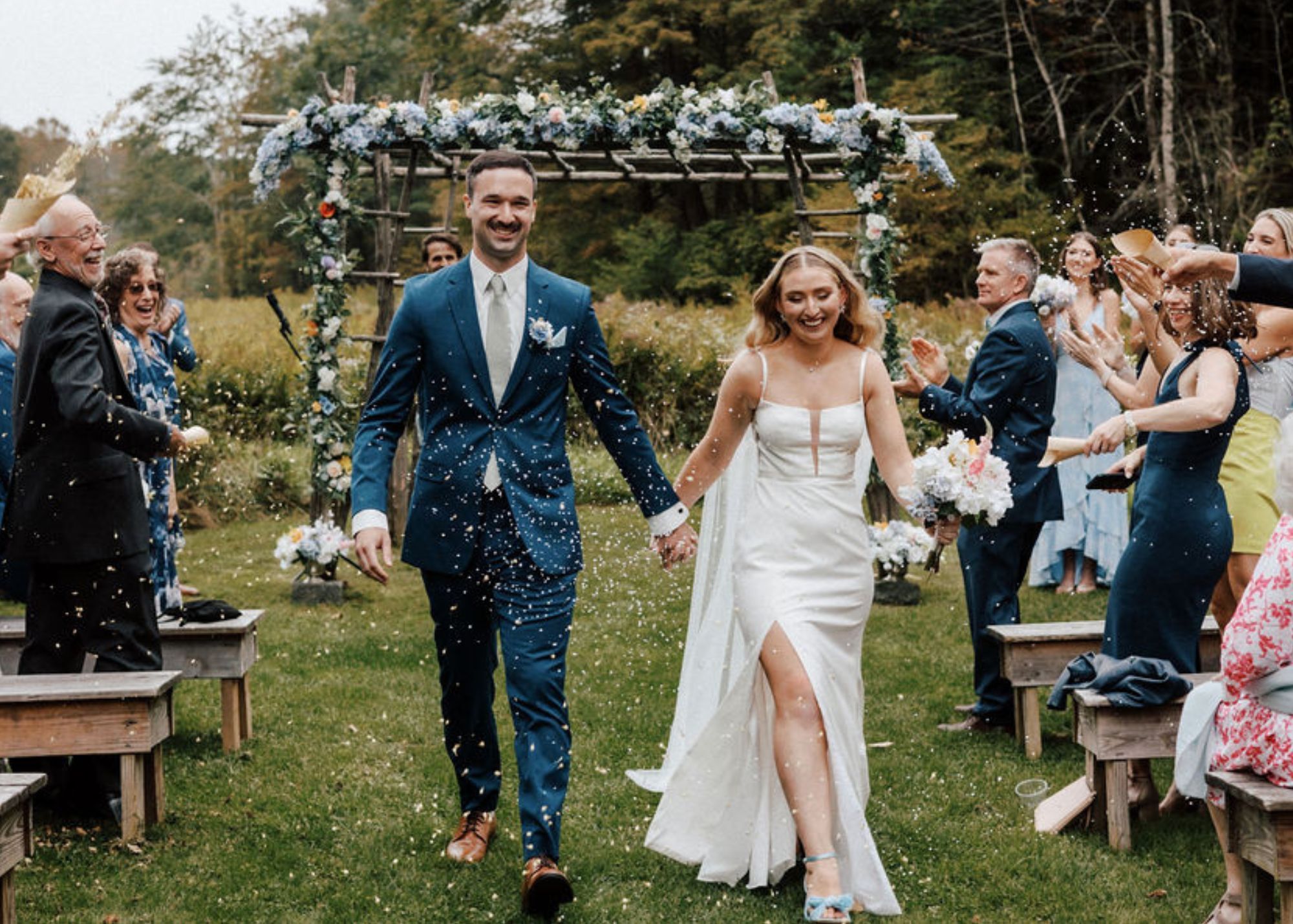 a bride and groom walk hand in hand up the aisle at their outdoor farm wedding. The bride wears a fitted wedding dress with a sheer cape veil. The groom wears a navy suit.