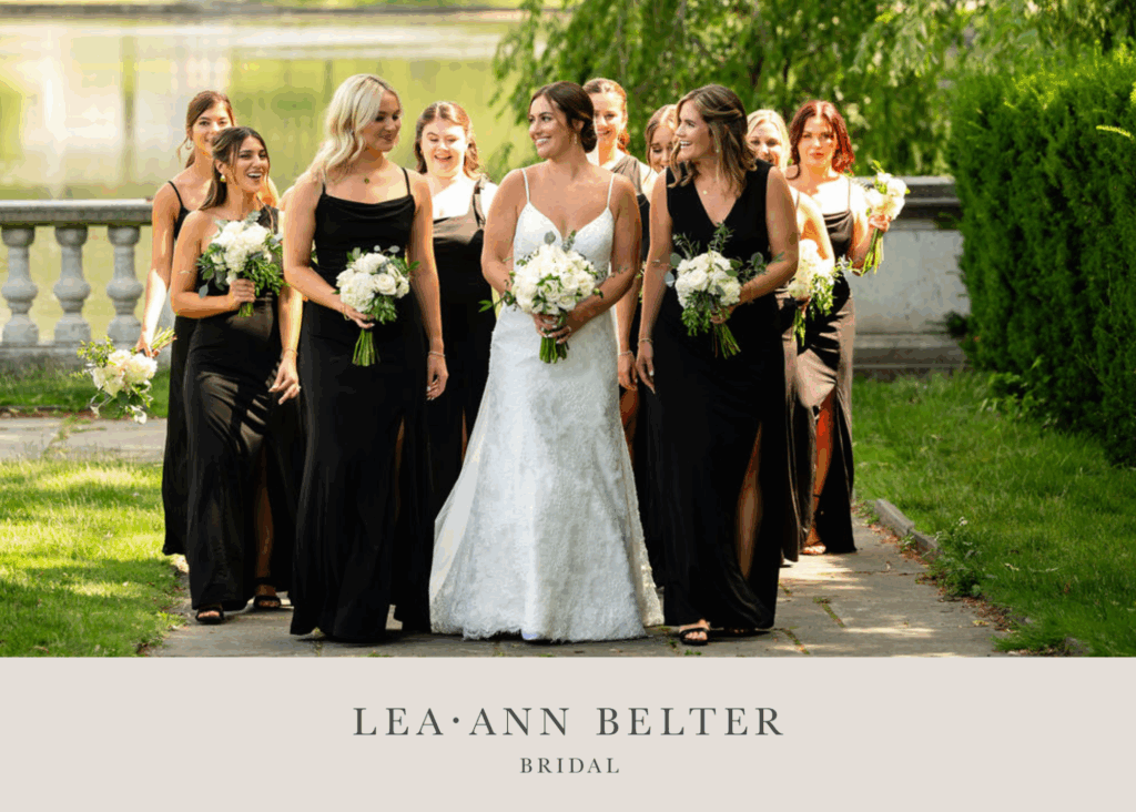 Bride wearing a lace wedding dress by Lea-Ann Belter Toronto walks with bridesmaids in wine dresses holding white bouquets on a sunny outdoor path.