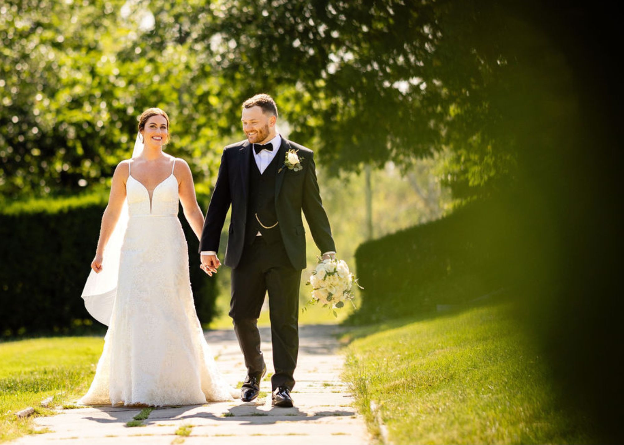 A bride and groom walk hand in hand in the sunshune. The bride wears a lace fit and flare with a cathedral veil